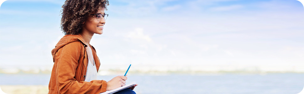 A smiling african-american woman outside, writing in a workbook. Reflecting on her thoughts.